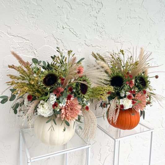 Fresh fall flower arrangements in white and orange pumpkins with cream and dusty pink blooms, sunflowers, greenery, and pampas grass.
