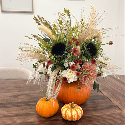 Orange pumpkin filled with fresh sunflowers, dusty pink and cream blooms, burgundy accents, greenery, and pampas grass.