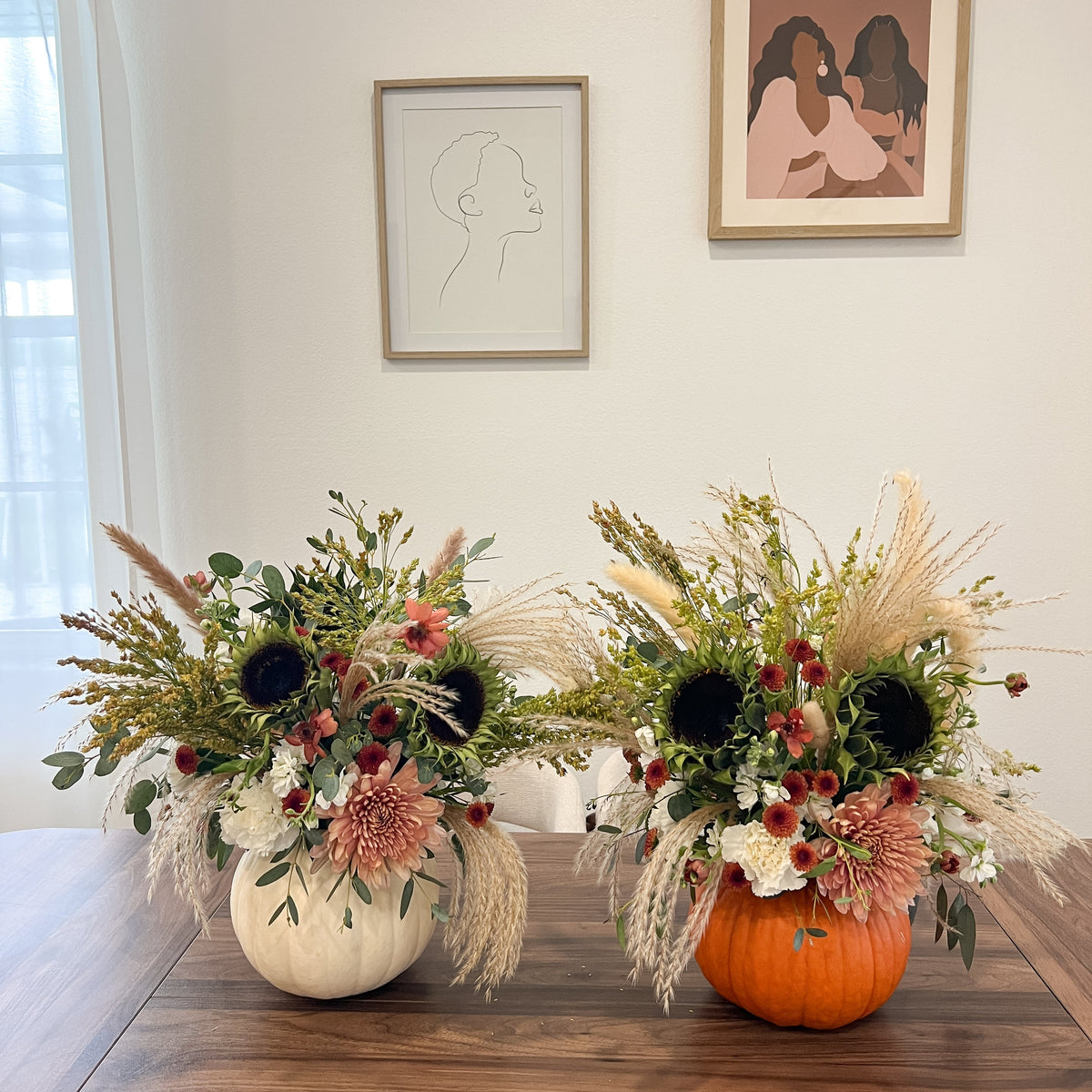 Fresh fall flower arrangements in white and orange pumpkins with cream and dusty pink blooms, sunflowers, greenery, and pampas grass.